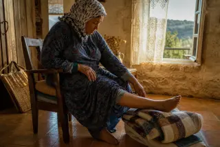 Senior woman stretching gently while seated in a warm, sunlit Charente living room, rustic décor, late afternoon ambiance, no artificial posing.