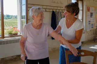 Two senior women in a bright village hall, one helping the other stretch near a chair, relaxed and smiling with rural details and vineyards visible through the window.