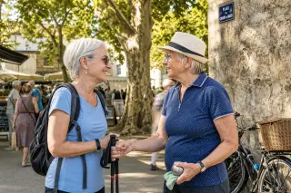 Two senior women greeting each other in a lively village square near Angoulême, with market stalls and bicycles in the sunlit background.